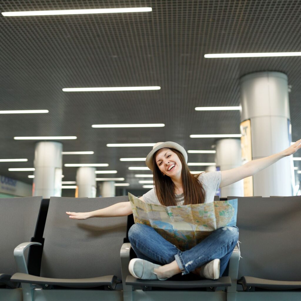young-happy-traveler-tourist-woman-with-paper-map-sit-with-crossed-legs-spreading-hands-as-flight-wait-lobby-hall-airport